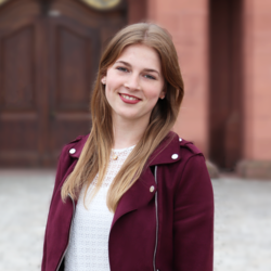 Sara Klein. Sie hat braune lange Haare und trägt eine weiße Bluse mit roter Jacke und steht vorm Mannheimer Schloss.