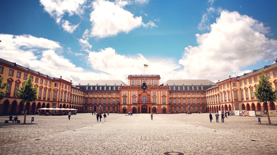 Das Barock-Schloss und der Ehrenhof der Universität Mannheim unter strahlend blauem Himmel. Das Schloss ist von unzähligen Fenstern, rotem Sandstein und einer gelben Fassade geprägt.