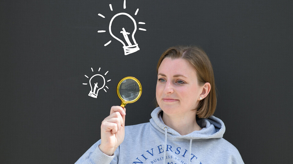 Junge Frau im grauen Hoodie hält eine Lupe vor gezeichneter Glühbirne auf dunklem Hintergrund.