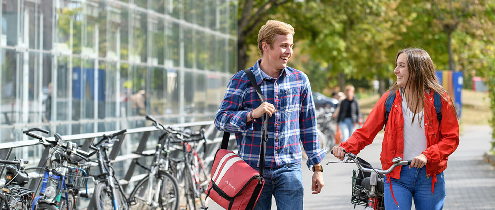 Ein Student und eine Studentin mit einem Fahrrad laufen zusammen und lachen sich an. Auf der linken Seite von ihnen sind viele Fahrräder abgestellt.