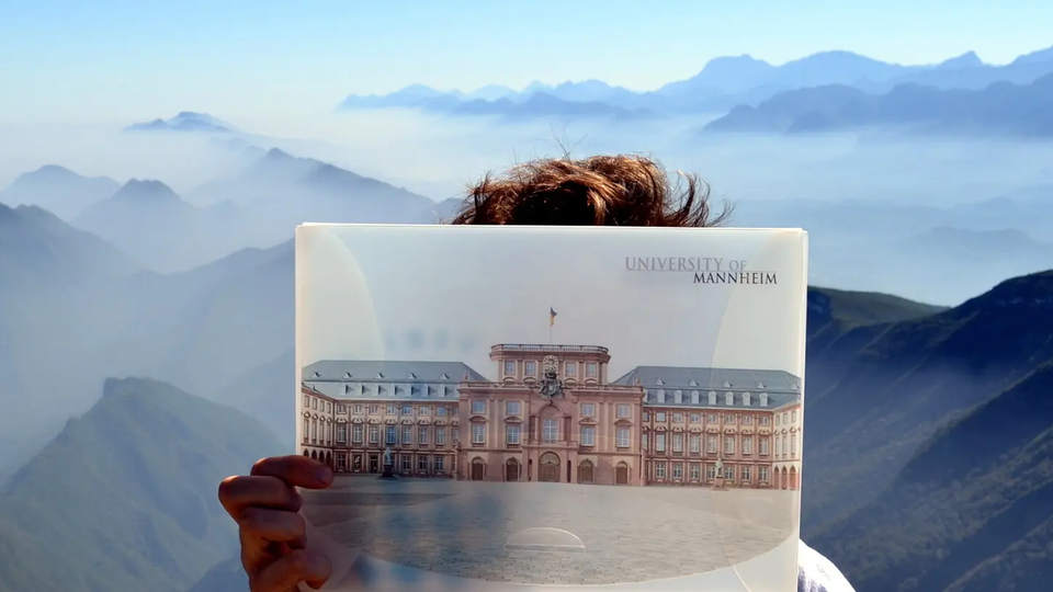 Person hält Foto des Mannheimer Schlosses vor Bergkulisse mit Nebel und Fernsicht in den Hintergrund.