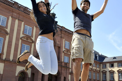zwei Studenten (links Frau, rechts Mann), die vor dem Schloss Uni Mannheim glücklich in die Luft springen