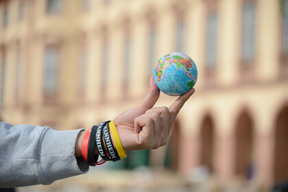 Hintergrund: Schloss Uni Mannheim, Vordergrund: ein Arm ragt ins Bild, zwiscen den Fingern wird ein Mini-Globus präsentiert