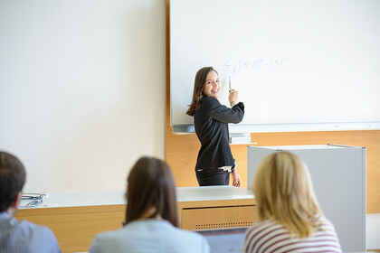 Lehrende an der Tafel und Studierende von hinten im Hörsaal