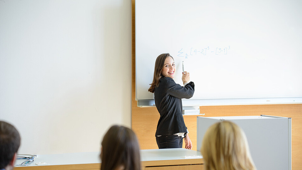 Lehrende an der Tafel und Studierende von hinten im Hörsaal