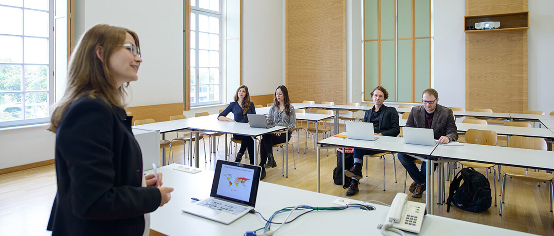 Eine Dozentin steht gegenüber von vier Studierenden, die teilweise mit dem Laptop am Tisch sitzen.