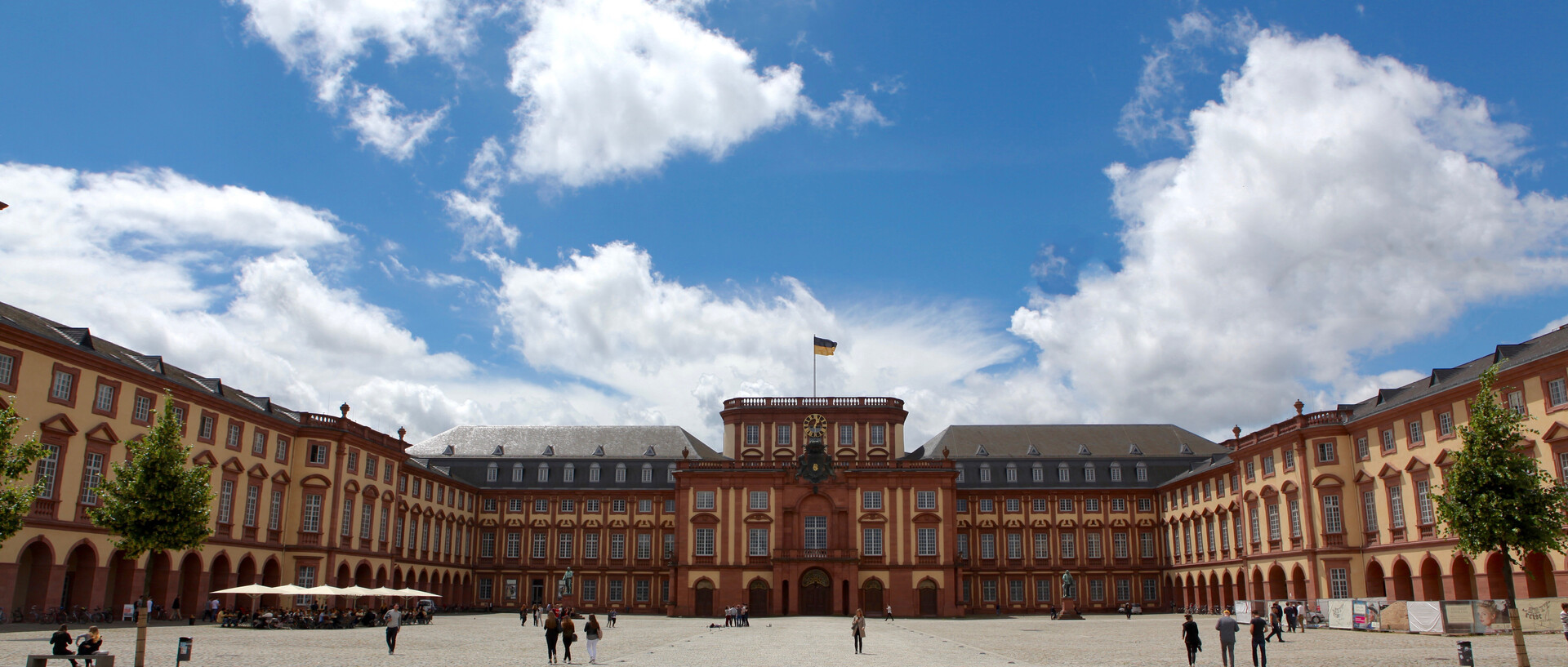 Das sandsteinfarbene Mannheimer Barockschloss mit Blick auf den Mittelbau bei strahlend blauem Himmel. Vereinzelt schlendern junge Menschen über das Kopfsteinpflaster auf dem Ehrenhof.