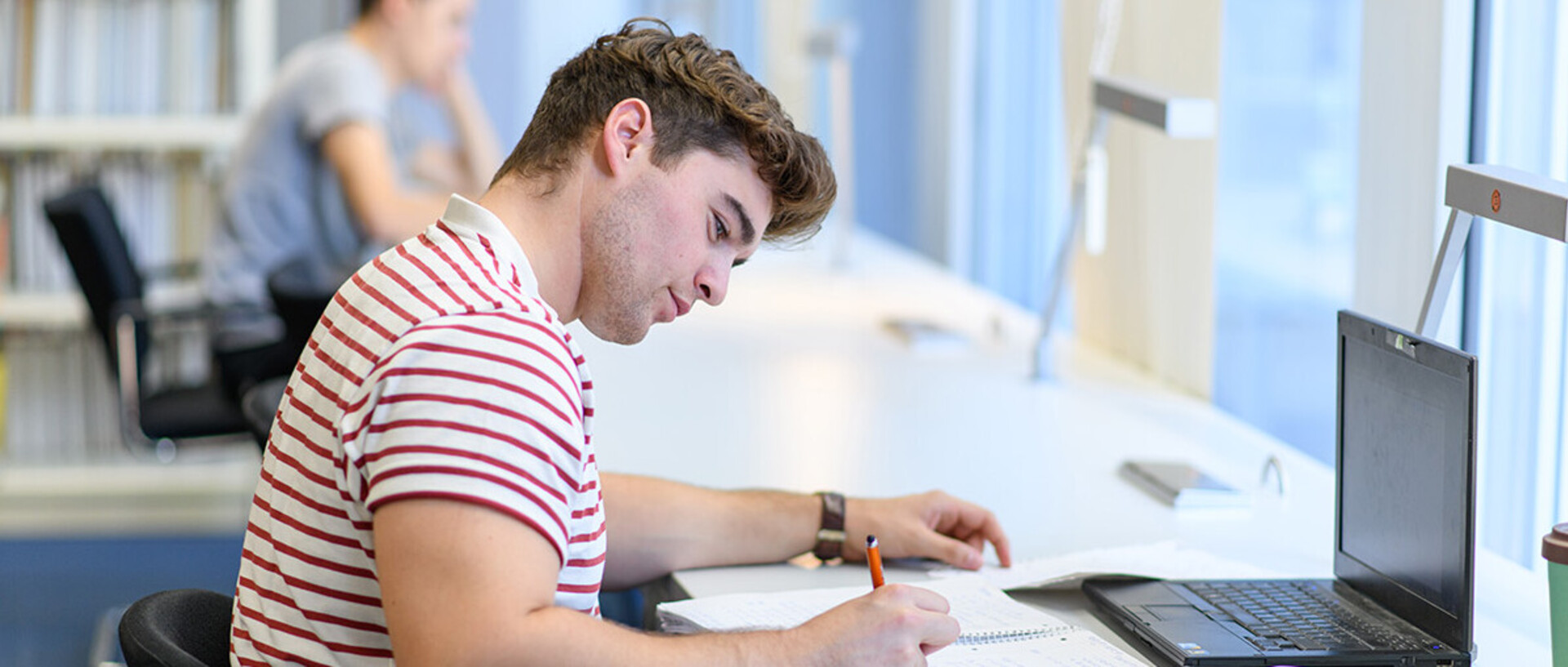 A student is sitting in the library in front of his laptop and writing something on a piece of paper.