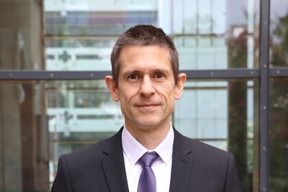 Martin Schlather hat kurze braune Haare. Er trägt einen schwarzen Anzug und eine lilane Krawatte.