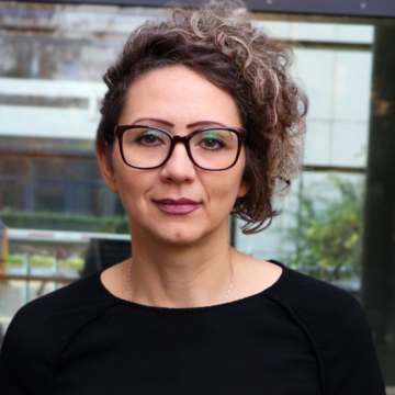 Sanja Juric hat dunkle lockige kurze Haare. Sie trägt eine schwarzumrandete Brille und ein schwarzes Oberteil.