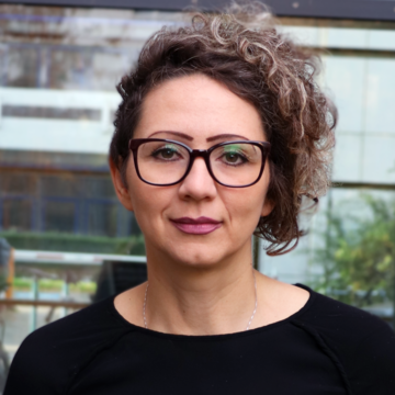 Sanja Juric hat dunkle lockige kurze Haare. Sie trägt eine schwarzumrandete Brille und ein schwarzes Oberteil.