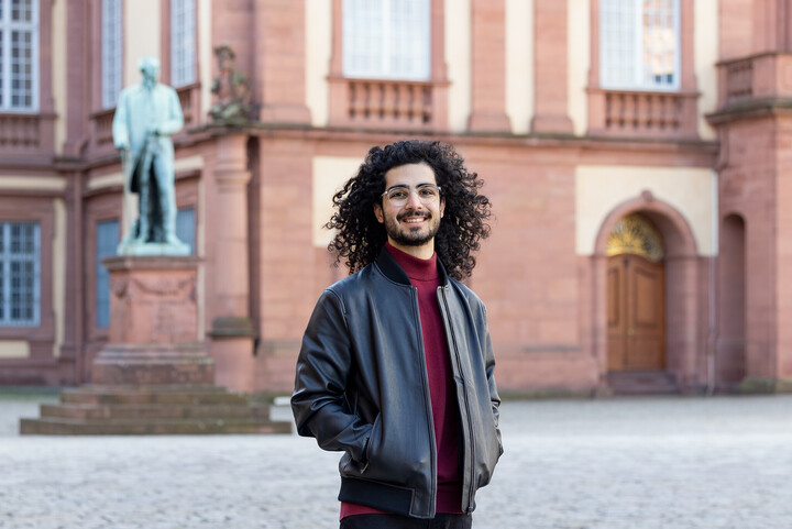 Eliya Ameri hat offene lockige schwarze Haare und lächelt vor dem Schloss in die Kamera. Er trägt eine Brille und eine Lederjacke.