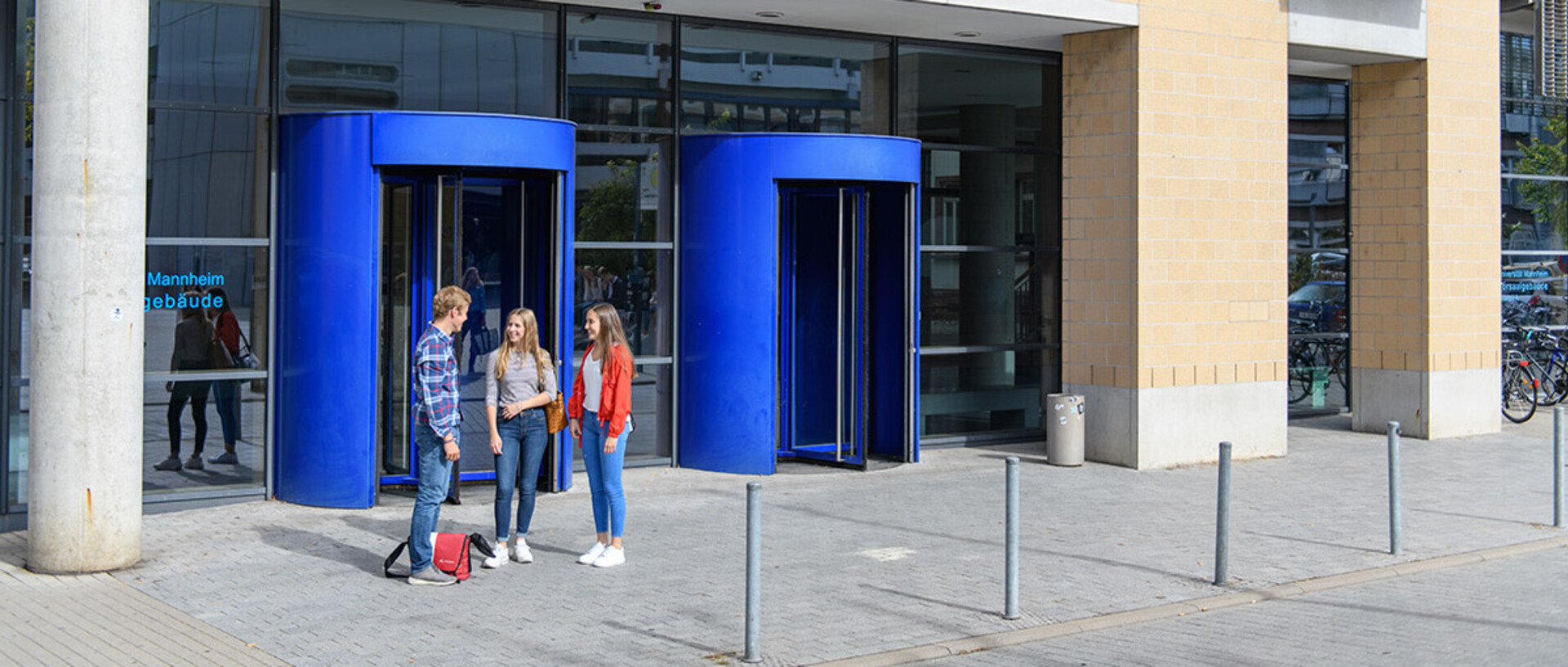 Three students are standing in front of the revolving doors in B6, talking to each other.