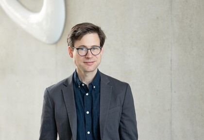 Portait von Prof. Staudigl: Junger Mann mit Anzug und Brille steht vor einer Wand der Fakultät für Wirtschaftsinrofmatik und Wirtschaftsmathematik