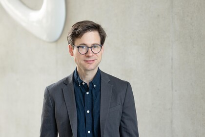 Portait von Prof. Staudigl: Junger Mann mit Anzug und Brille steht vor einer Wand der Fakultät für Wirtschaftsinrofmatik und Wirtschaftsmathematik