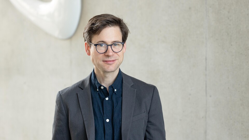 Portait von Prof. Staudigl: Junger Mann mit Anzug und Brille steht vor einer Wand der Fakultät für Wirtschaftsinrofmatik und Wirtschaftsmathematik