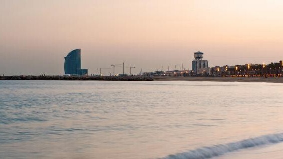 Strandpanorama bei Sonnenuntergang mit ruhigem Meer, Barcelonas Skyline und dem markanten W-Hotel im Hintergrund.