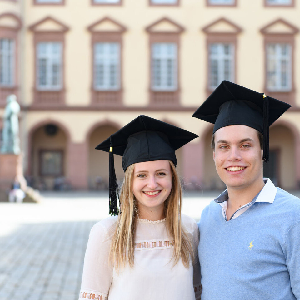 Eine Bachelorabsolventin und ein Bachelorabsolvent stehen vor dem Mannheimer Schloss.