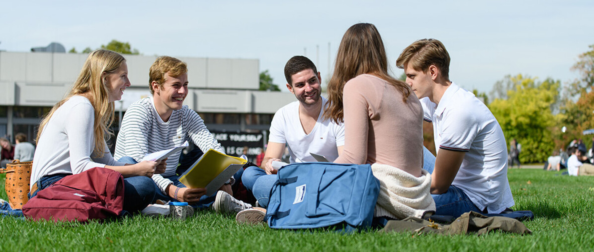 Five students are sitting in a circle on a lawn, laughing.