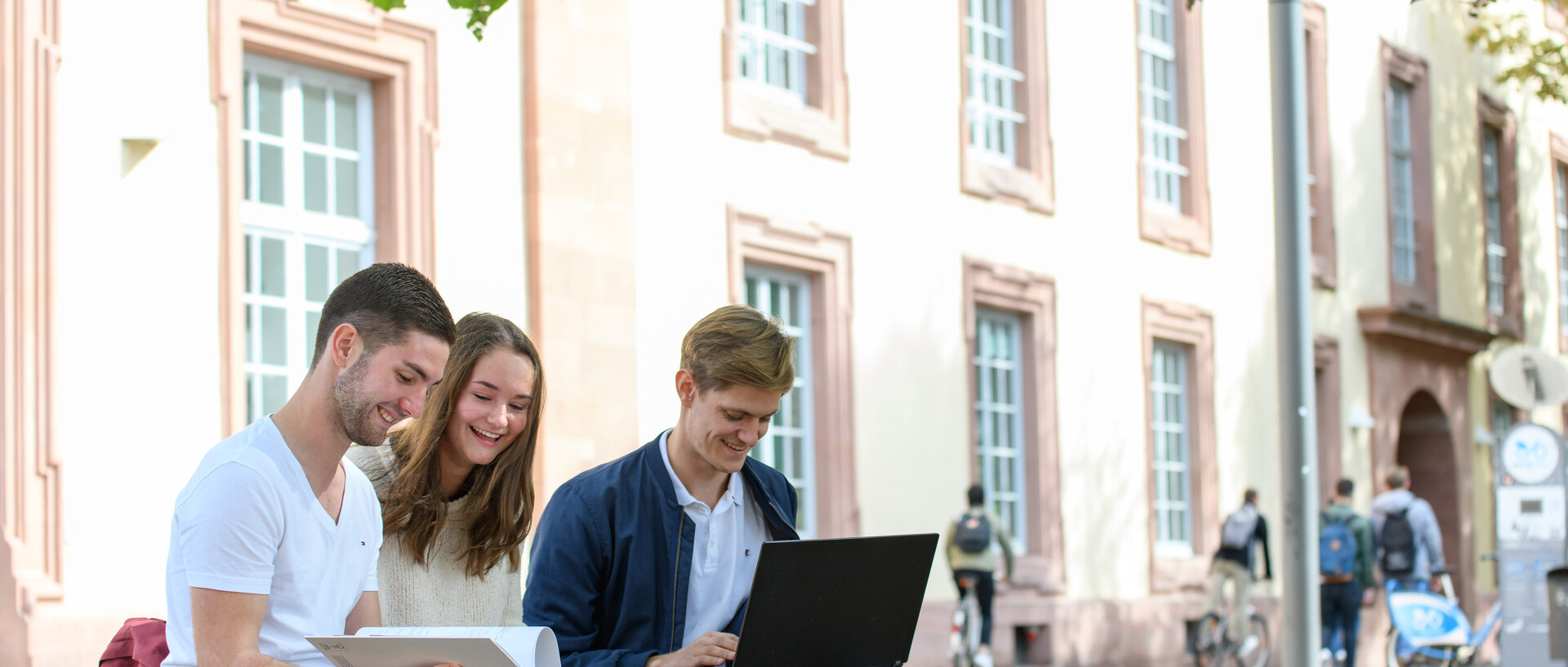 Three happy students with learning materials sitting in front of the university