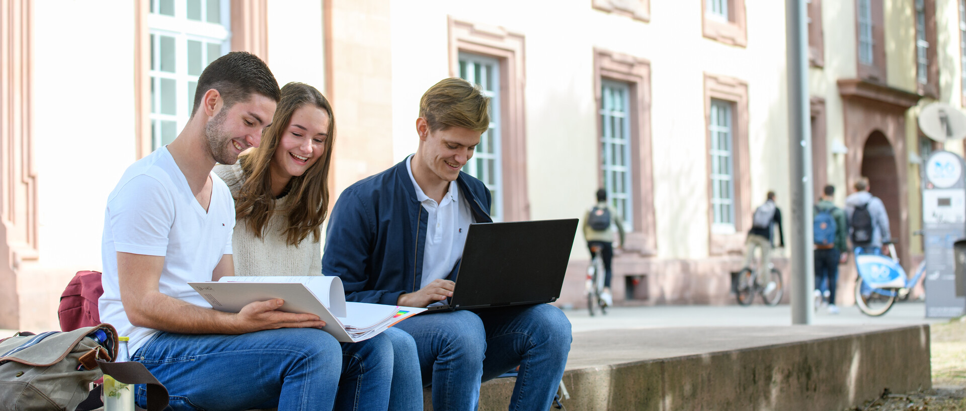 Drei Studierende sitzen vor der Universität und schauen in einen Ordner und Laptop